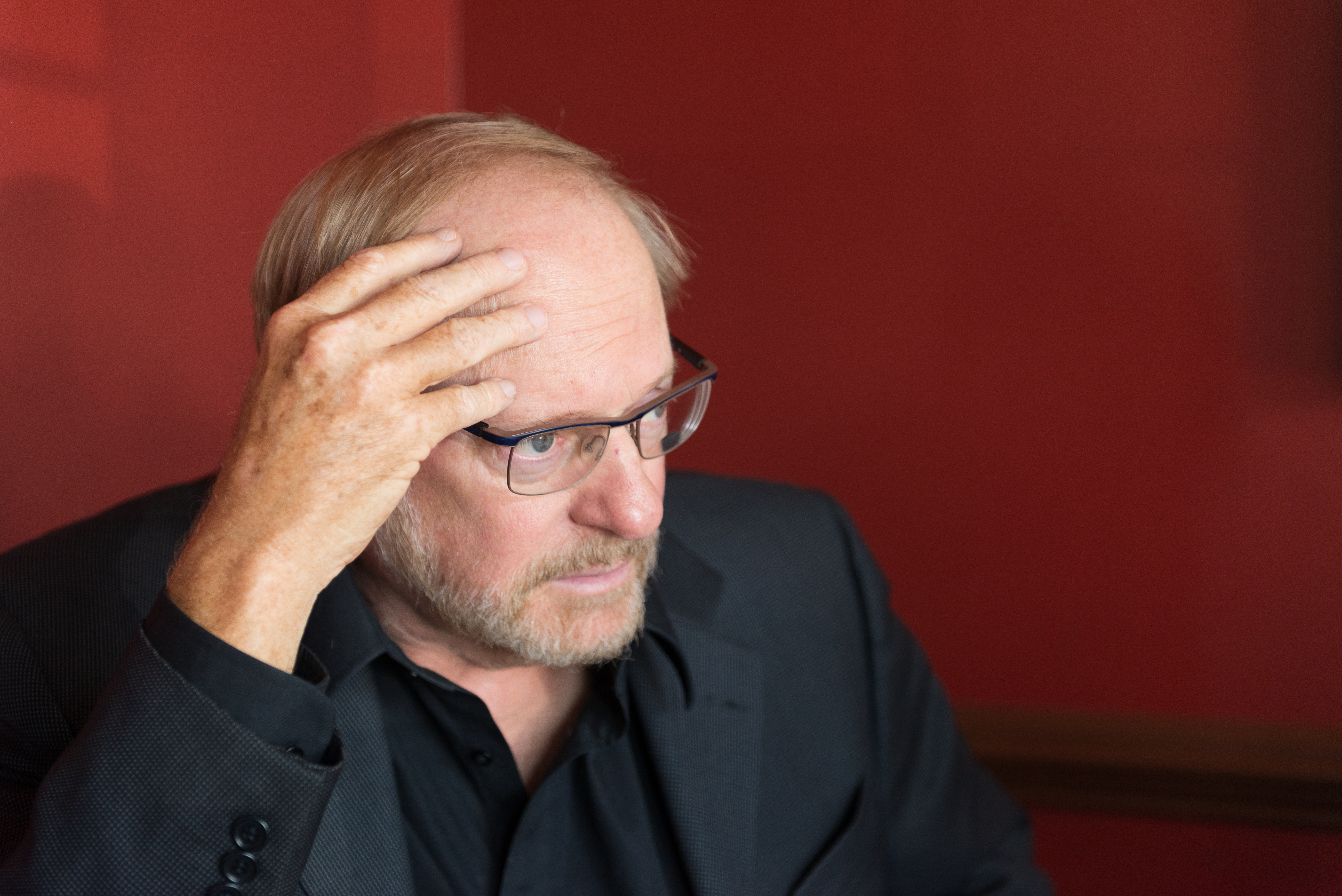 Medium close-up of George Gopen, a white man with short reddish greying hair, mustache and beard. He has blue eyes and rectangular glasses with metal frames, and is looking pensively to the right, while his open left hand touches on his forehead, as if deep in thought. He is wearing a black collared shirt and dark grey jacket. To his right, are three individual images of books he has written. The background is a deep maroon color.