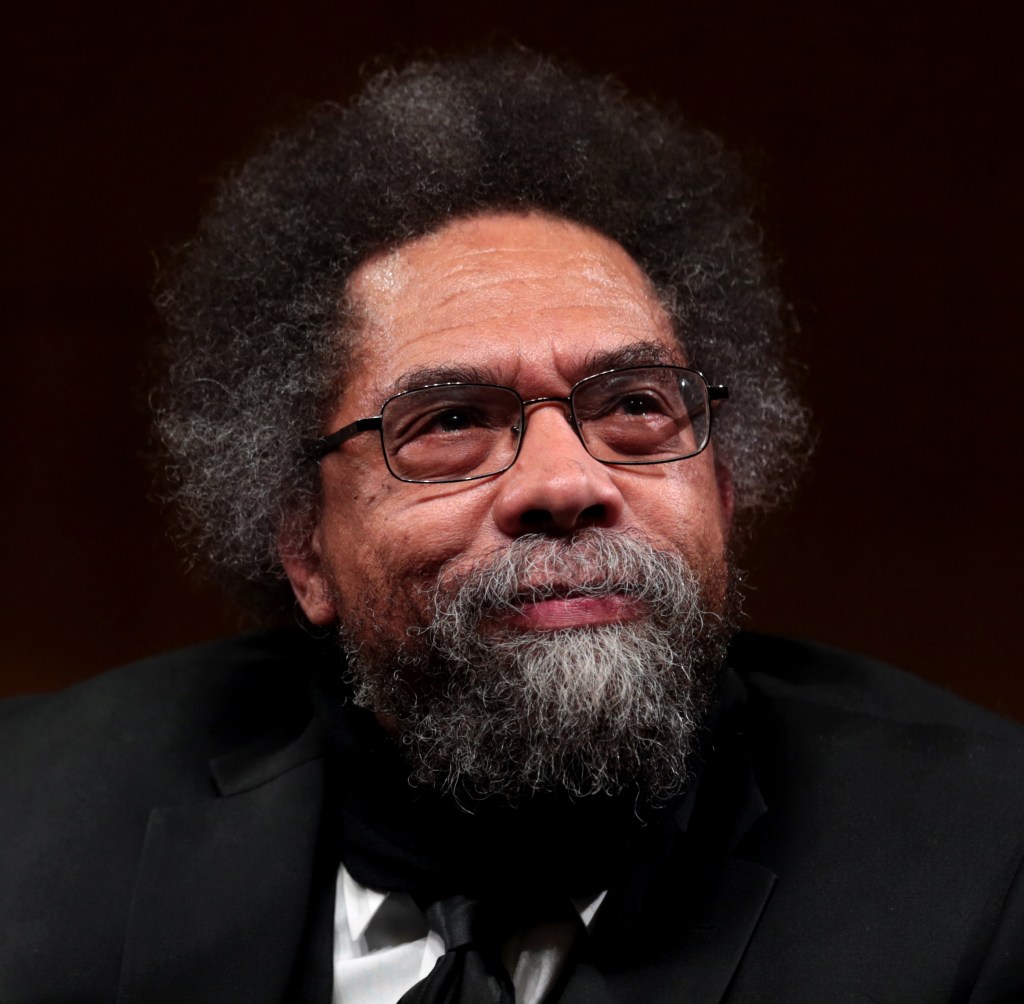 Head shot of Cornel West, a smiling Black man with black and grey afro, beard, and mustache, tinted glasses in a black jacket and white shirt with a small microphone pinned to it.
